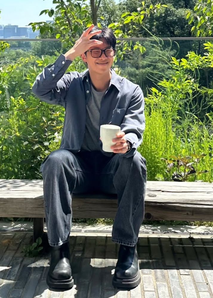 photo of smiley glasses namjoon sitting out in the sun with a cup, moving the hair away from his forehead. 