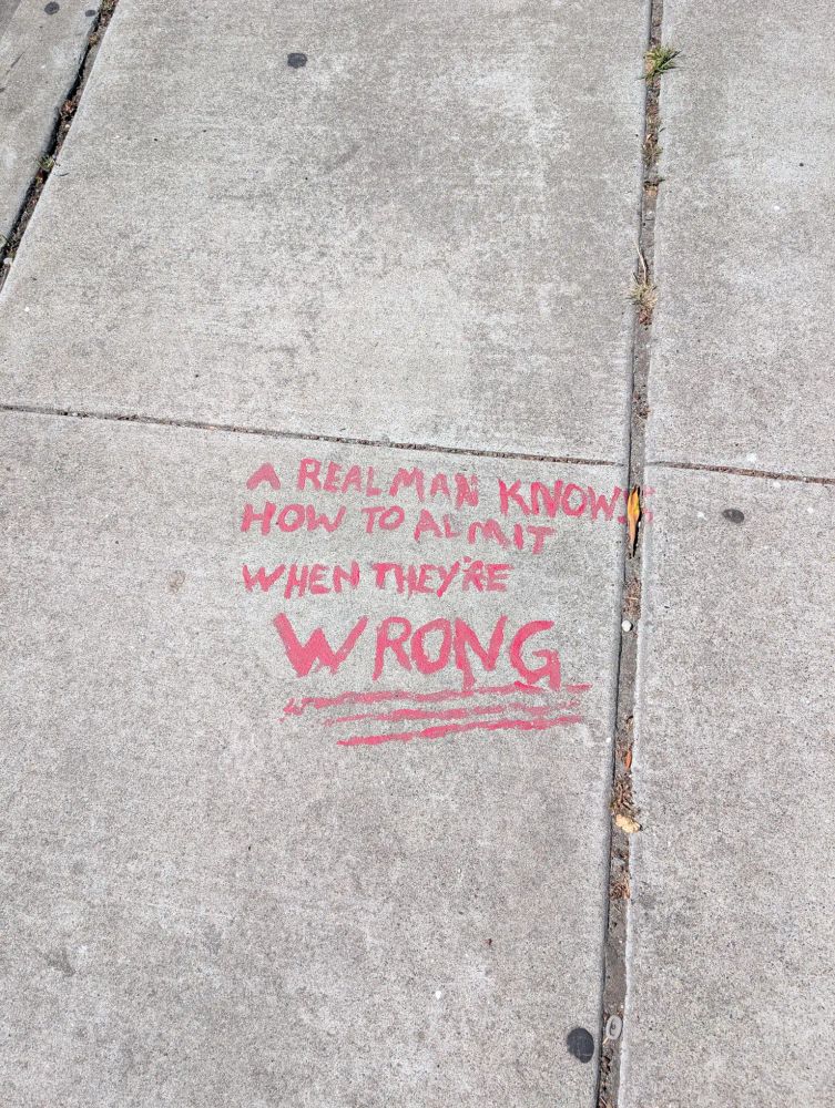 Sidewalk graffiti in red reading "A real man knows how to admit when they're wrong"