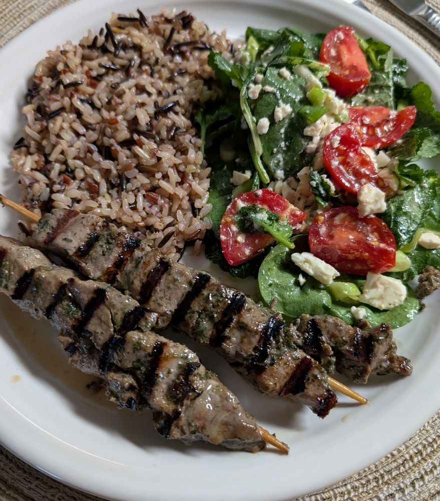Wild rice blend top left, salad top right and two grilled lamb skewers at the bottom on a round white plate.