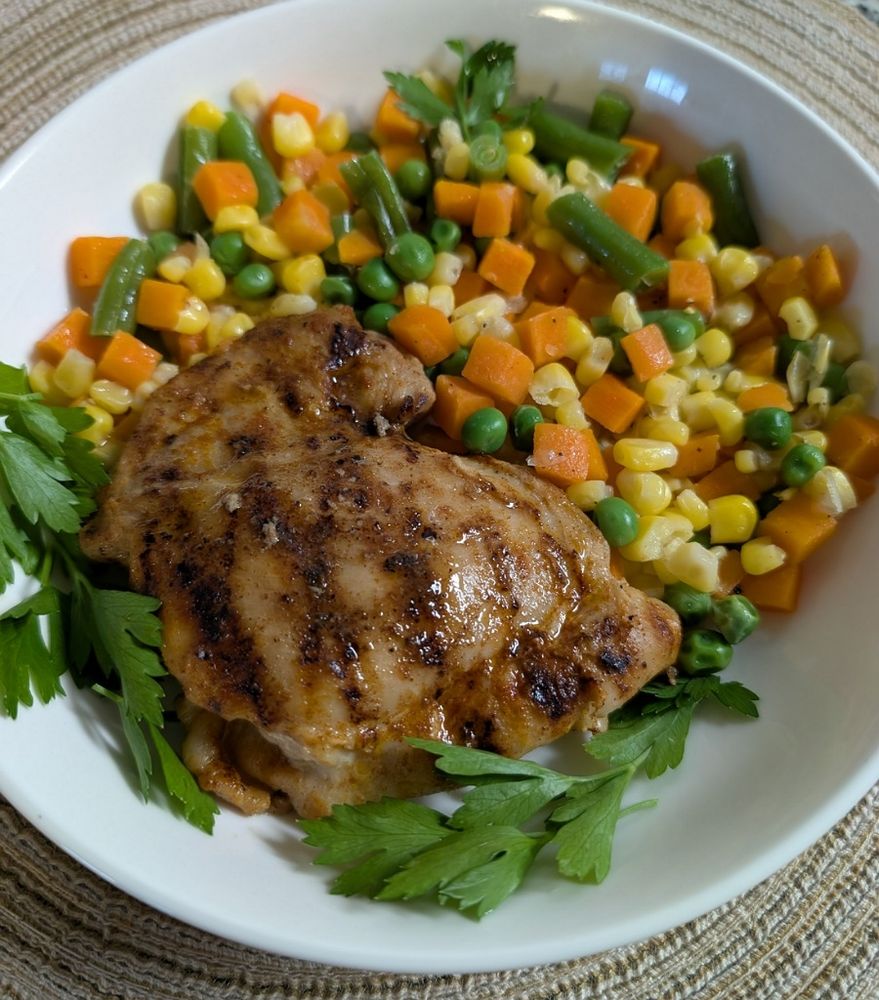 The dinner described the text in a round white bowl on top of a round brown placemat. The veggies are at the top and the chicken thighs at the bottom surrounded by some parsley.