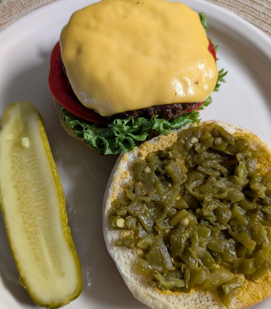 At the top of a round white plate is the bottom portion of the hatch chili cheeseburger, with mustard, onion, lettuce, tomato and the cheeseburger patty. Bottom right is the top half of the bun with mustard and chopped Hatch chili. And bottom left is a dill pickle half.