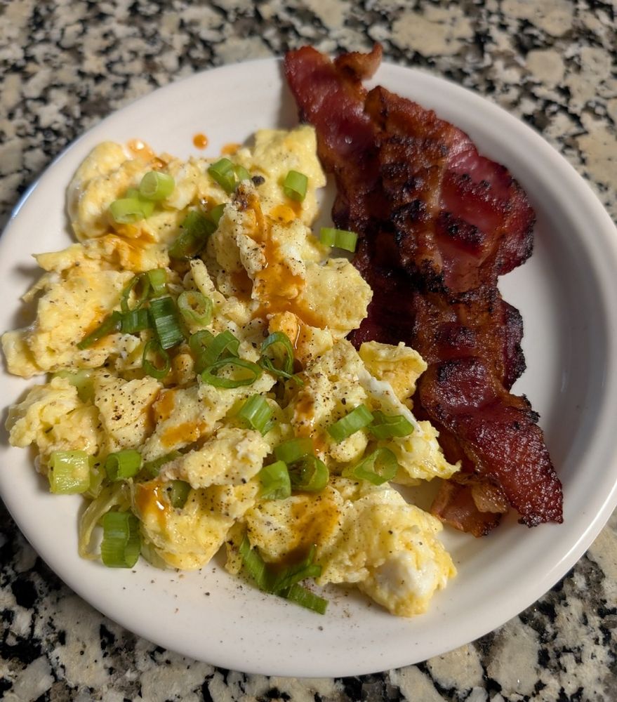 Scrambled eggs with cheese and green onion and Cholula sauce and two strips of thick bacon on a round white plate.