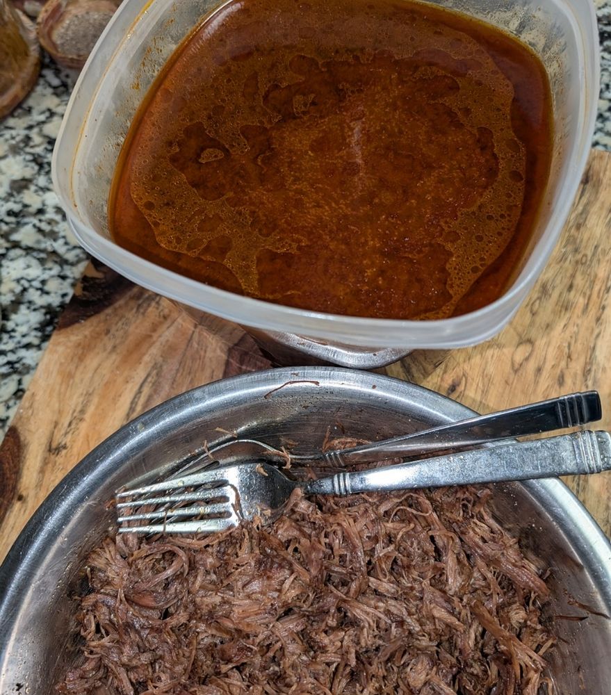 A tupperware container with the beef birria broth. Below is a a metal bowl with the shredded beef chuck roast.