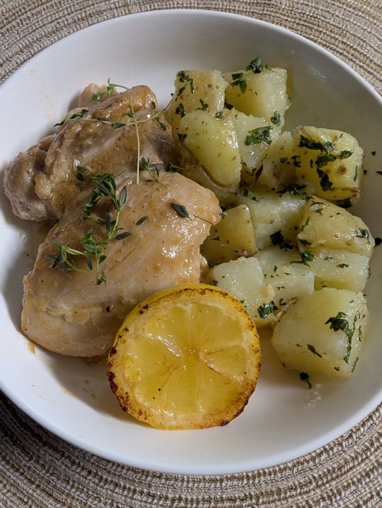In a round white bowl on top of a round light brown placemat is a sauteed chicken thigh on the left and parsley potatoes on the right and a sauteed wedge of lemon at the bottom.