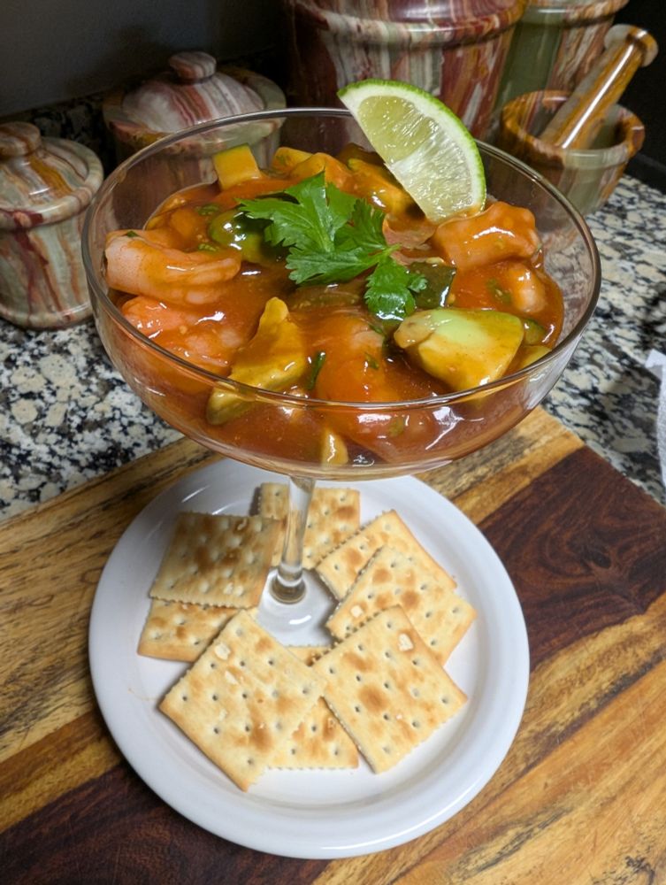 A Mexican shrimp cocktail in a margarita glass on a round white plate that has saltine crackers around the bottom of the glass.