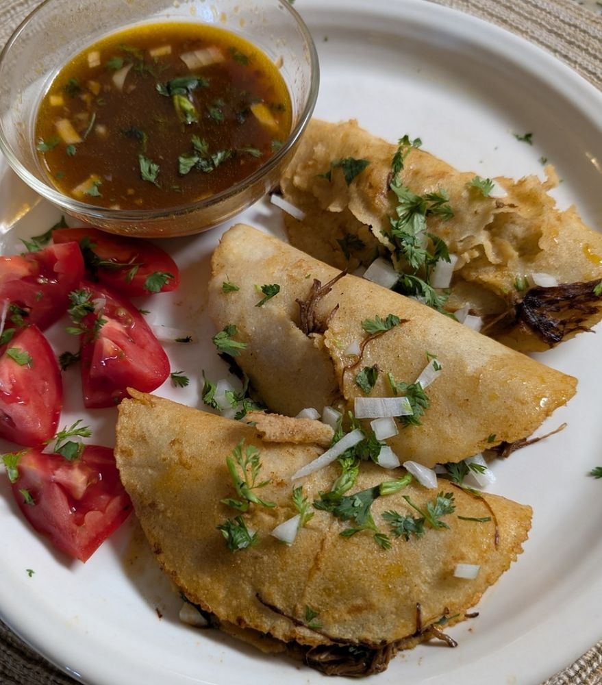 On a round white plank is the finished dish of three birria tacos on the right, tomatoe wedges on the bottom left and in the upper left some of the beef broth for dipping.