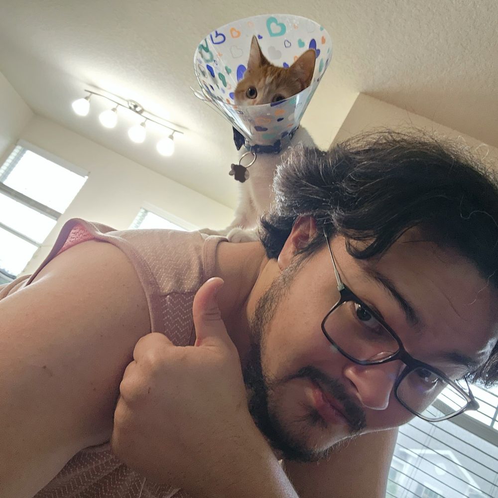 An Orange Tabby kitten (approximately 4 months in age) wearing a post surgery cone is standing on a man's shoulder looking into the distance. The man gives a reassuring thumbs up, even though his back is dying.