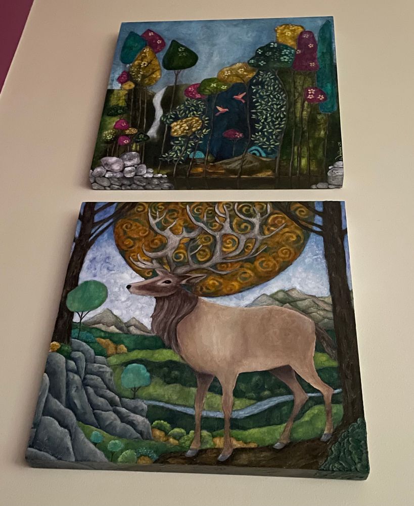 Two paintings hanging on a cream coloured wall. The bottom one is of a male deer standing on a hill, a large swirly sun behind it, with mountains, boulders, and trees in the background. The top one depicts a fantasy landscape with multicoloured trees, a waterfall, and little birds. 