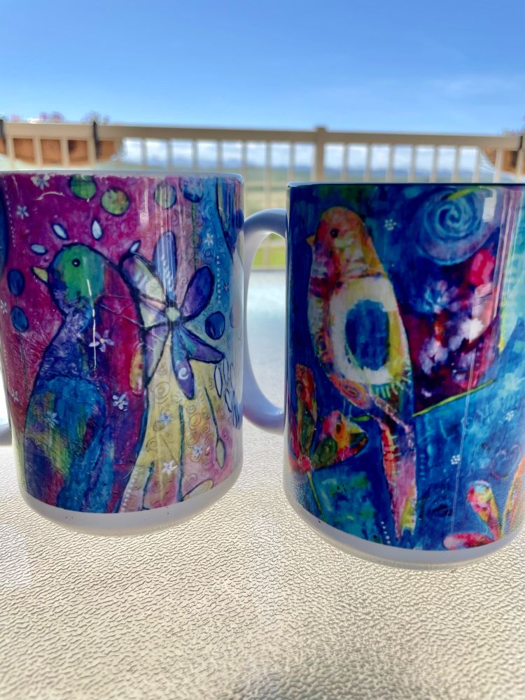 Two colourful art mugs with birds and flowers in them sit on a patio table. 