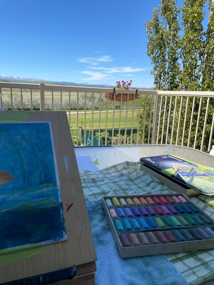 An art easel with a pastel work in progress sits next to a colourful array of soft pastels on a table on a deck with a view of mountains in the distance. 