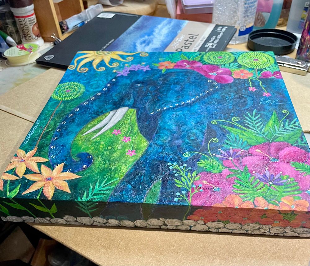 Colourful acrylic painting sits on a table. It features a blue elephant walking through a garden of pinwheel trees, bright magenta yellow and peach tropical flowers, and various bright green plants. It wears a crown of small purple flowers. There is a swirly sun in the upper left corner. 
The bottom of the canvas is painted to continue the scene, with a line of white stones at the elephant's feet.