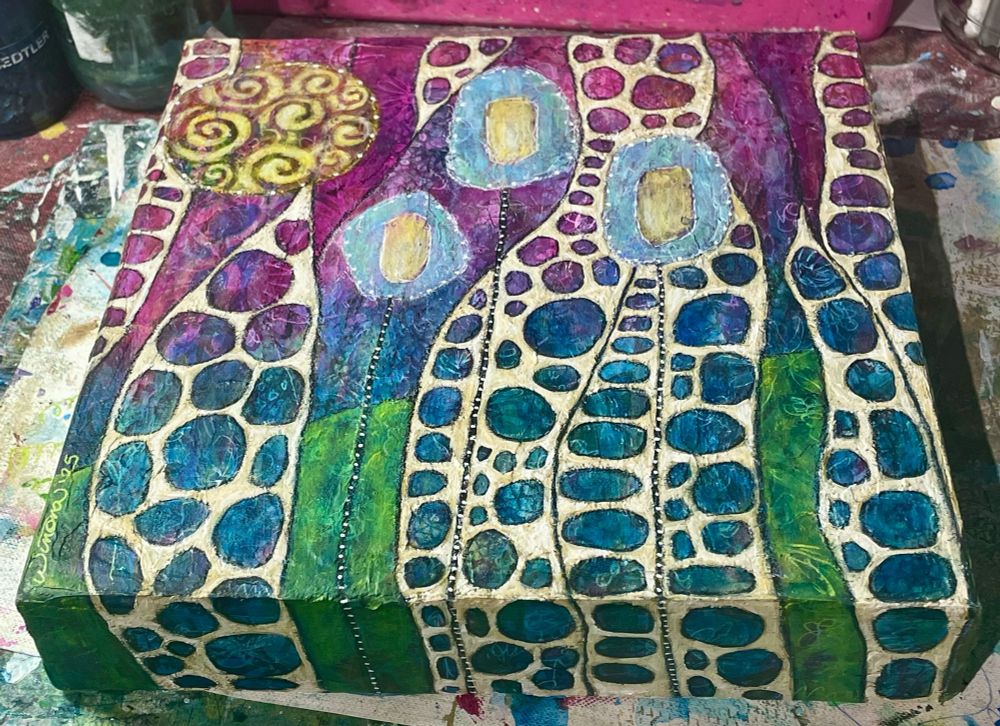 Vividly coloured acrylic painting called "Infinity Ribbons" featuring lots of circles in blues and pinks on tan coloured ribbon-like shapes, with a swirly sun in the upper left and three circle shaped blue trees in the centre. The land area is bright green and the sky area is bright magenta. 