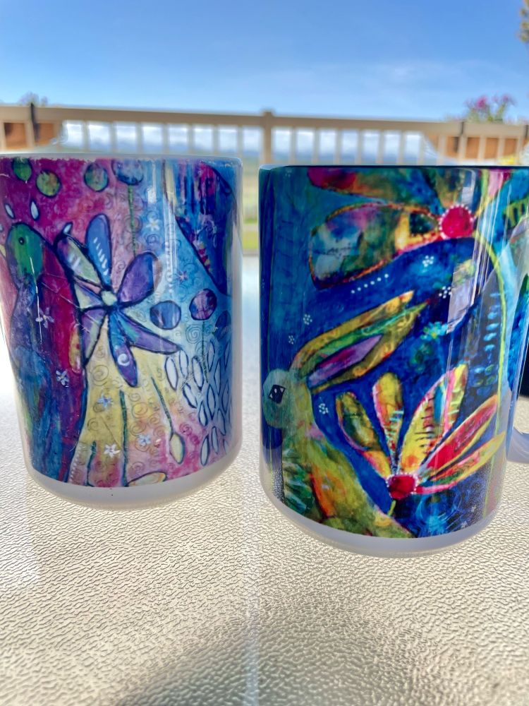 Two colourful art mugs with birds and flowers in them sit on a patio table. 