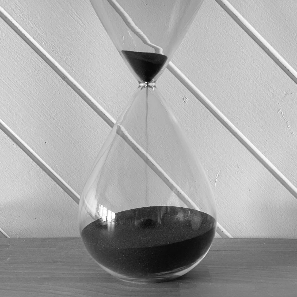 A black and white photograph of an hourglass on a wooden surface, with a white diagonally panelled wooden wall behind it. The sand in the hourglass is black. Most of the sand is already in the bottom half, leaving only a small amount still to fall. 