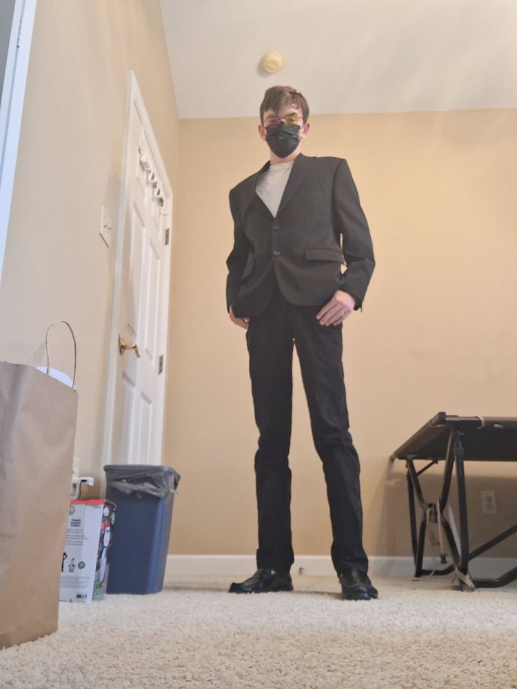 tall teen dressed rather in a white shirt, black jacket, black pants, and back shoes. the really noticeable thing about the wear are the pink and yellow glasses, resembling Spamton from Deltarune 