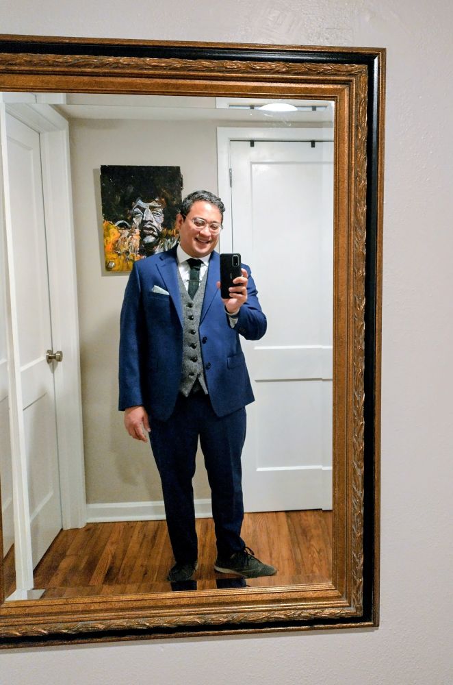 Guy in blue suit and grey vest in a standard American apartment.
