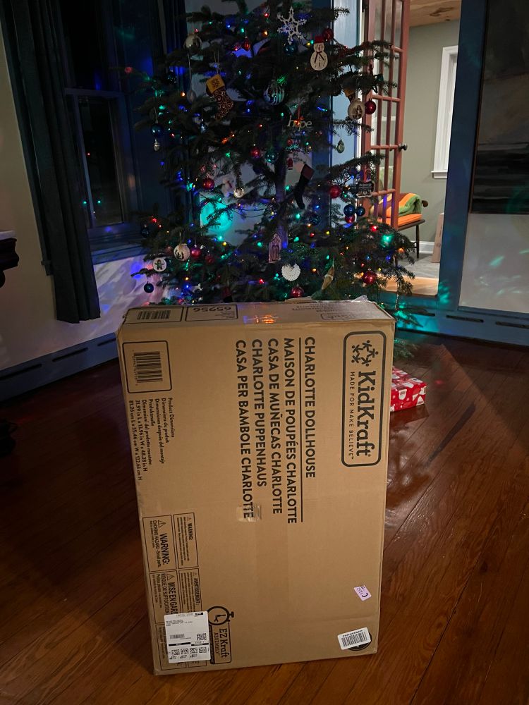 Large cardboard box in the foreground, Christmas tree in the background. The box says KidKraft Charlotte Dollhouse.