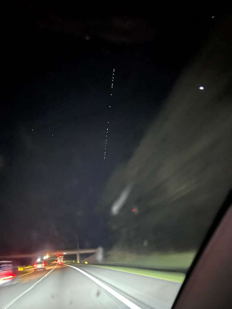 Highway at night as seen through car windshield. Above the highway, in the sky, is a pin-straight string of lights.