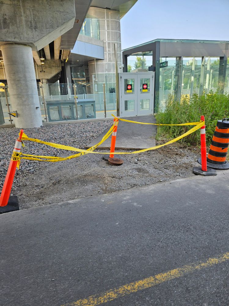 The same area as the previous asphalt volcano, now with pavement removed, and a trio of orange plastic construction posts with a festive yellow caution tape connecting them, surrounding the graveled space. 