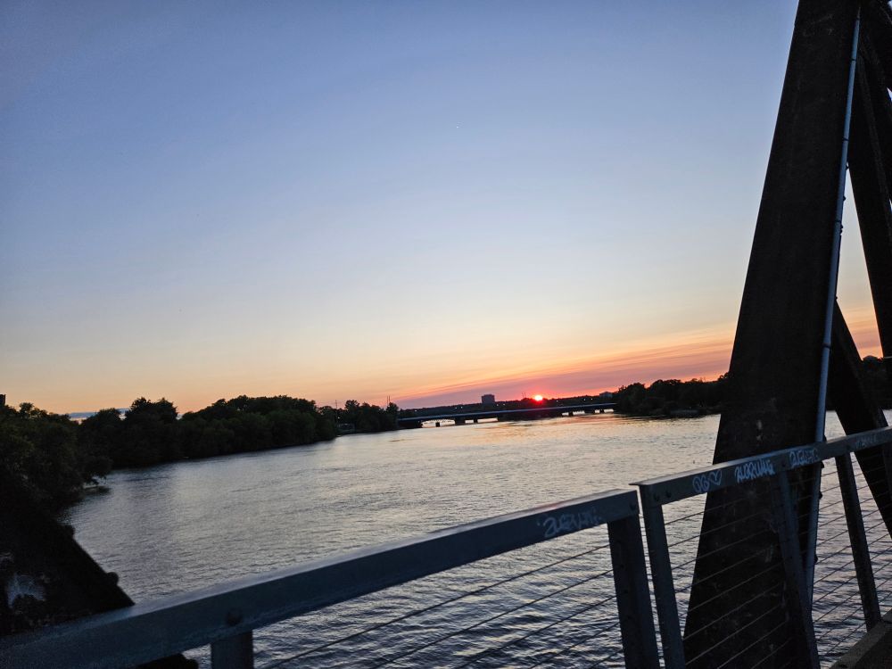 The sun can be seen just dipping below the horizon line over the Ottawa River. A bridge truss is at the right side of the frame. The photographer definitely should have been paying more attention to riding her bike than taking this photo, but alls well that ends well. 