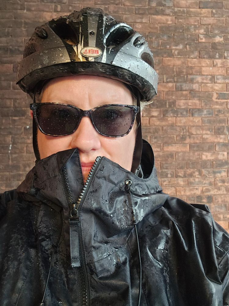 A person wearing a bike helmet, sunglasses, and a wet rain coat. 