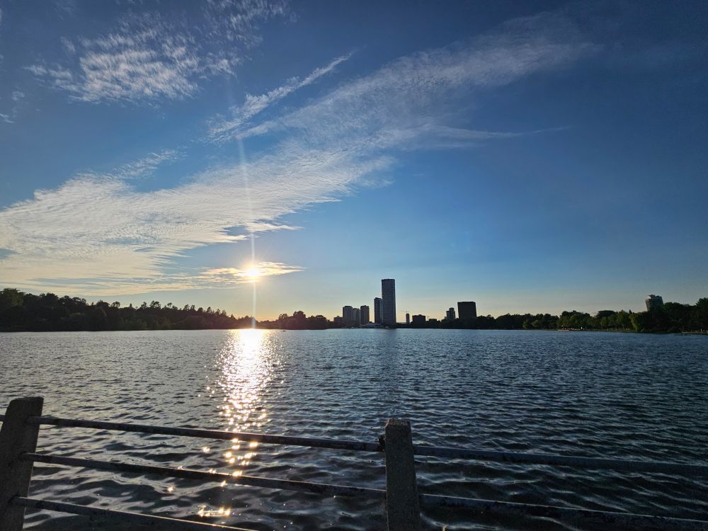 Image of the sun, low in a blue sky with scattered wispy clouds. The sun is shining on Dow's Lake, in Ottawa. A few buildings and an ascending line of trees are along the horizon. 