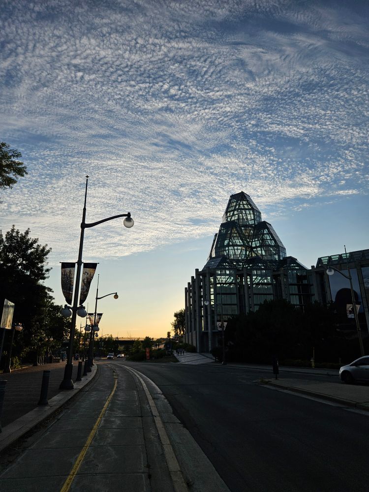 The glass dome of the National Art Gallery, and a lamppost, is silhouetted against a white cloud dappled blue sky at sunset. The image is taken from a bi-directional bike lane. 