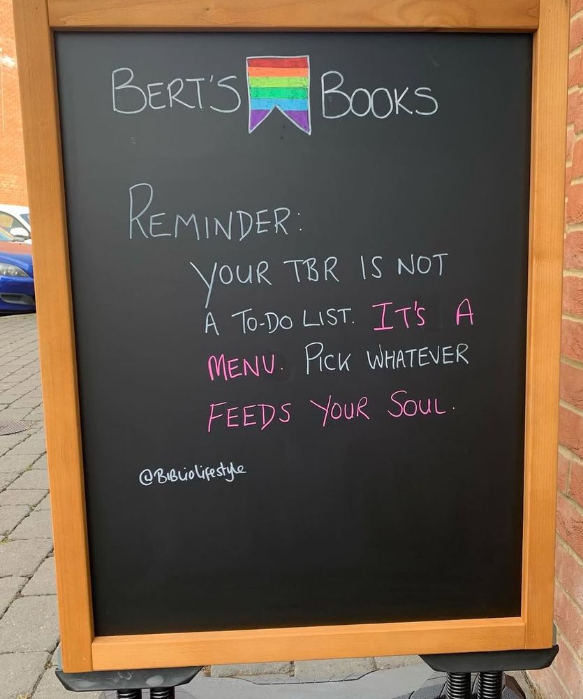 Alt text: A black sandwich board sign outside a bookstore named “Bert’s Books.” The sign features a hand-drawn rainbow-colored bookmark above the store name. The message reads:
“Reminder: Your TBR is not a to-do list. It’s a menu. Pick whatever feeds your soul.”
The quote is attributed to @bibliolifestyle, with select words highlighted in pink for emphasis.