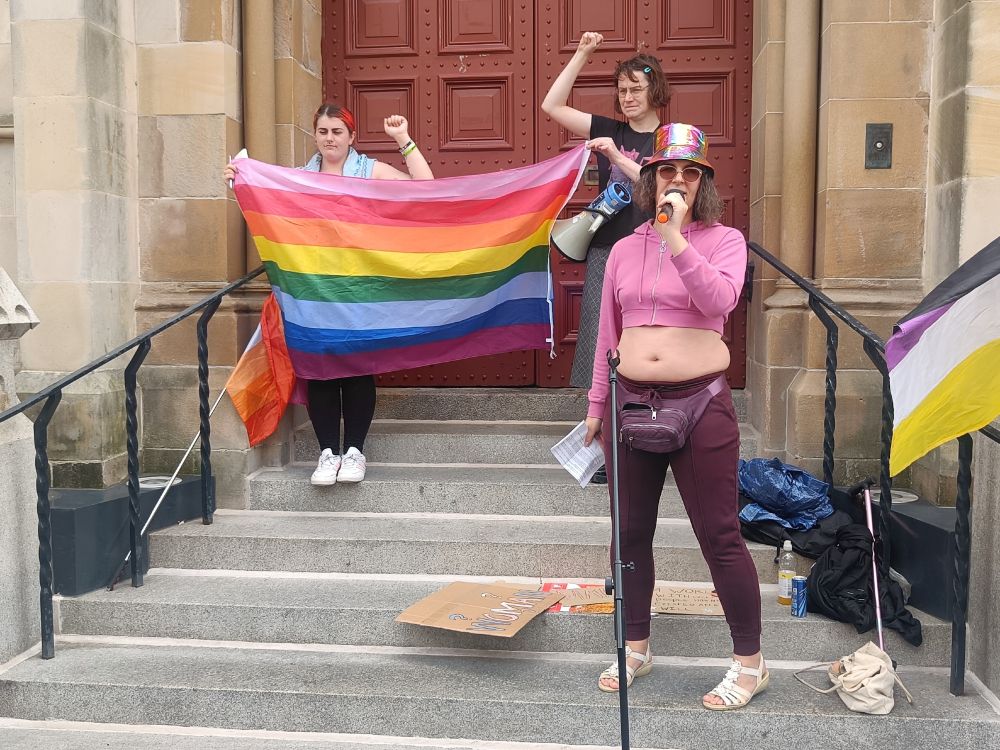 Toni speaks into the microphone on the town house steps. Sophie and Beth hold up an eight coloured rainbow flag behind Toni with fists in the air. 