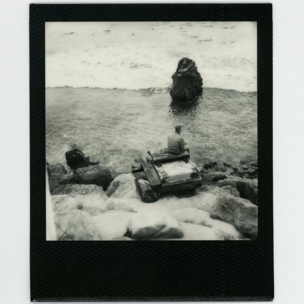 A black and white Polaroid image with a black frame. A man sits on the ruins of an old pier and stares out at the ocean. 