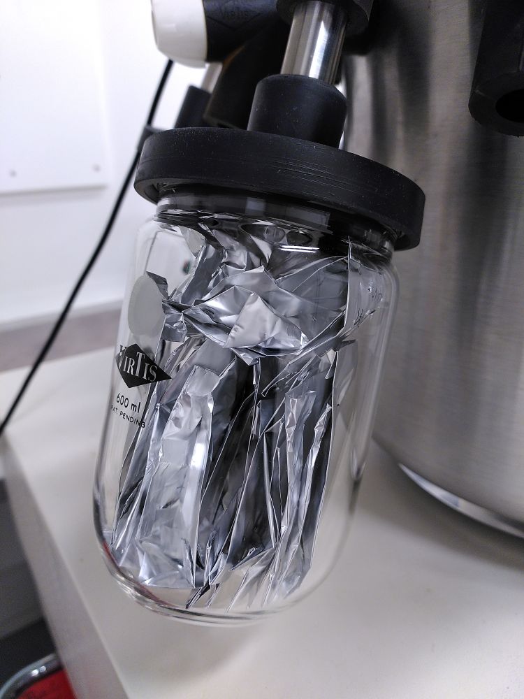 A close-up of a beaker from a freeze dryer with plant samples in aluminium bags. The glass beakers can be connected and the vacuum extends into the beakers with the sample in them. Black tubes connect the beakers with the vacuum chamber and white valves allow for individual connecting and disconnecting.