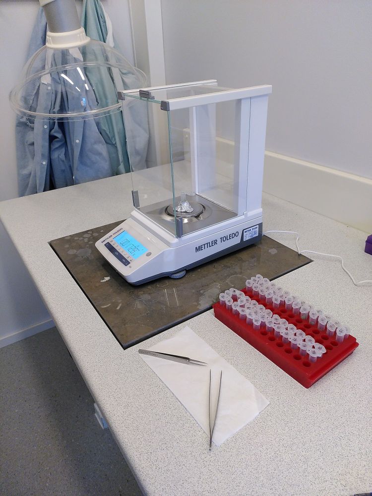 A fine scale for scaling in milligram range. The scale is resting on a special heavy table that prevents vibrations of the table to influence the scaling. A red rack with sample tubes is standing next to the scale as well as a clean tissue paper with two metal tweezers on it.