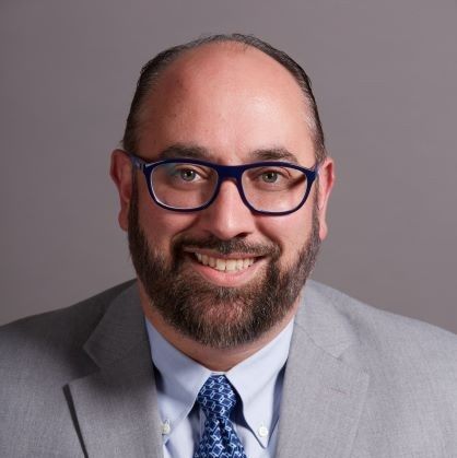 Headshot of a man with a beard wearing glasses, a suit jacket, and a patterned blue tie. He is smiling at the camera.