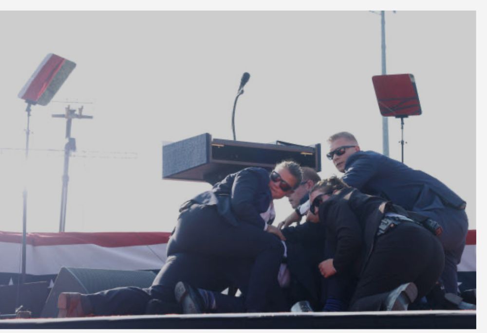 Men and women wearing dark suits are hunched down in a semi circle. Most are wearing dark sunglasses. All are seemingly looking toward a cameraman or director or other individual not seen in the photo. 
