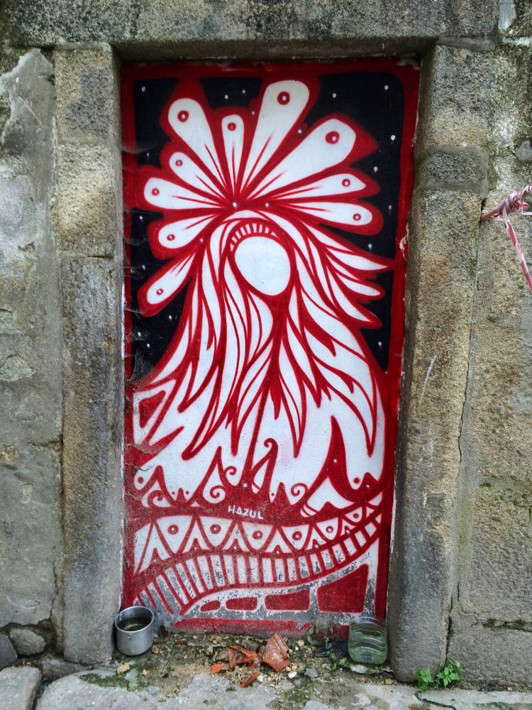 
A vibrant mural painted on a door inset in a stone wall. The mural depicts a stylized, otherworldly figure with a large, white head adorned with numerous swirling shapes. The figure's body is a deep red, with intricate patterns and lines. The figure has no face but is robed in a way which suggests a madonna or female saint. The background is a dark, starry night sky. The artist's signature, "HAZUL," is visible.

