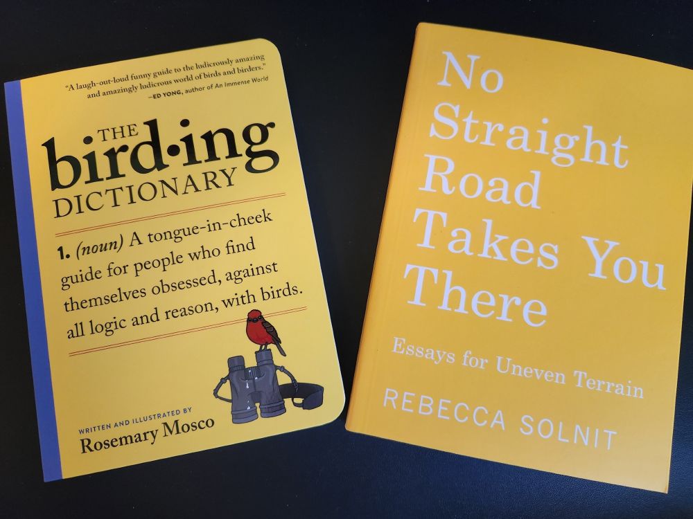 Two yellow books on a black background. The Birding Dictionary: A tongue-in-cheek guide for people who find themselves obsessed, against all logic and reason, with birds, written and illustrated by Rosemary Mosco. The cover is mostly yellow with a blue edge near the binding. There is an illustration of black binoculars with a bright red bird sitting on them. The second book is No Straight Road Takes You There: Essays of Uneven Terrain by Rebecca Solnit. It is bright yellow with white print. 