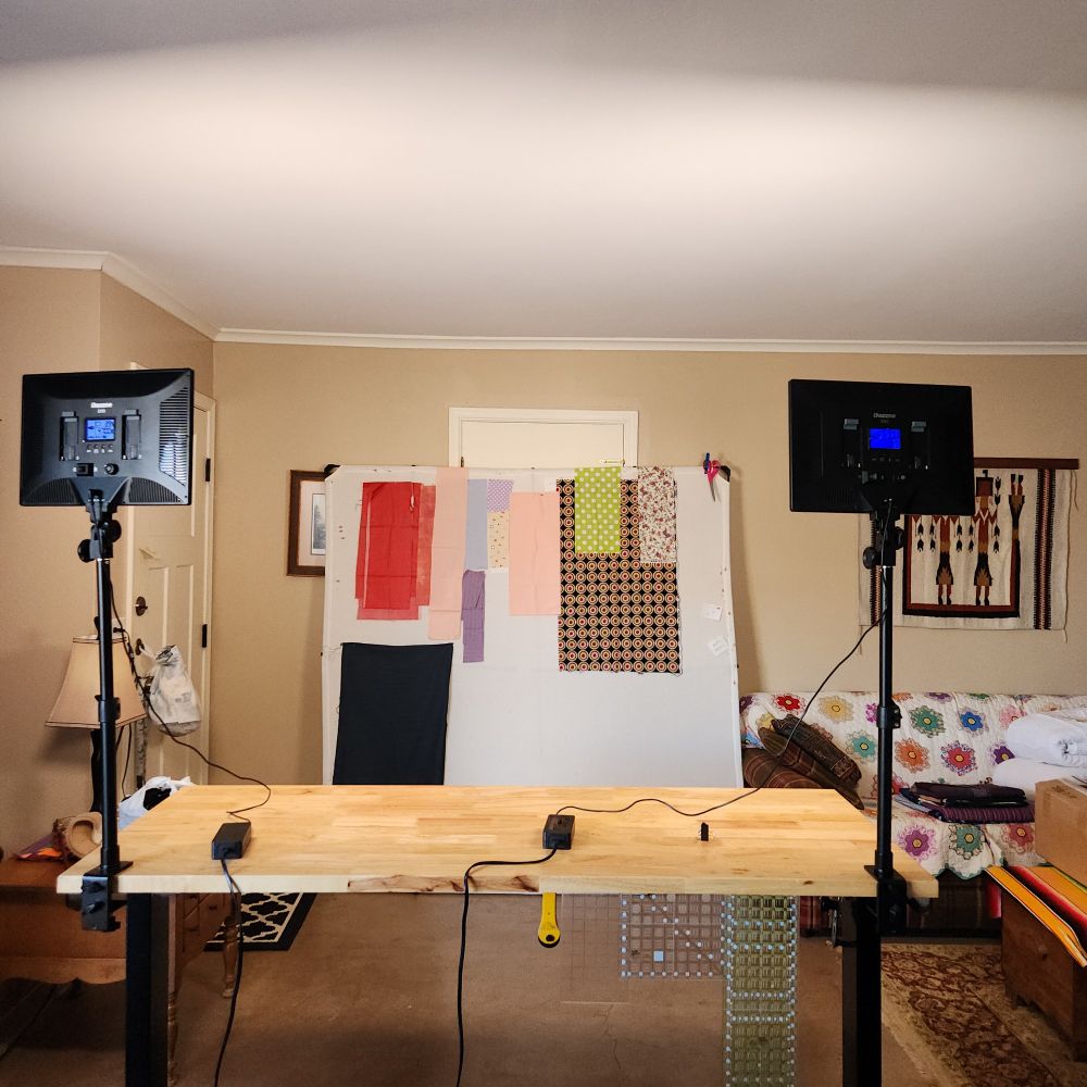 photo of Sarah's studio, aka living room. in the foreground a wood table is visible with two studio lights attached to it. in the background is a white flannel design wall, with various pieces of cotton fabric stuck on it.