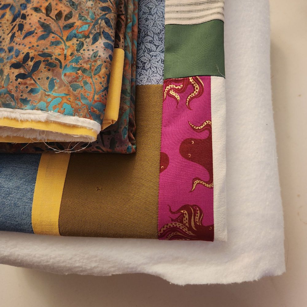 a pile made of two bundles of folded-up fabric on top of a fluffy white quilt batting. the stack is ready to take to the longarm quilter, but for now, is sitting on my ironing table. 