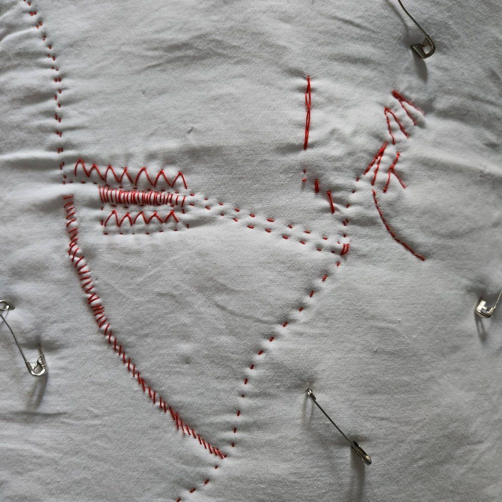 close up of a white pillowcase with various stitches in red thread. safety pins are holding the fabric layers together. 