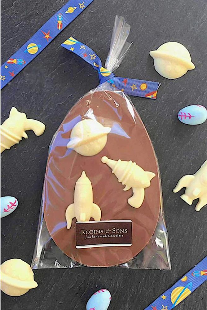 Space theme chocolate Easter egg bar. Novelty egg shaped milk chocolate topped with white chocolate space shaped, including planets, rayguns, rockets. In clear cellophane packaging tied with a blue multicoloured space themed ribbon.