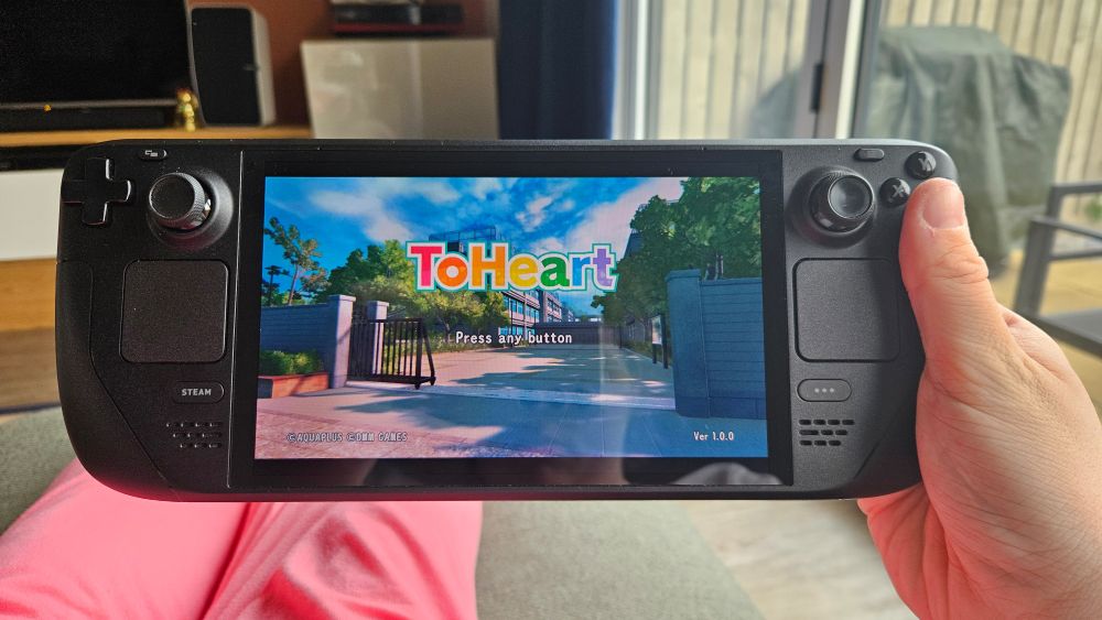 Milla is lying on a sofa, with the ToHeart remake title screen visible on her Steam Deck.