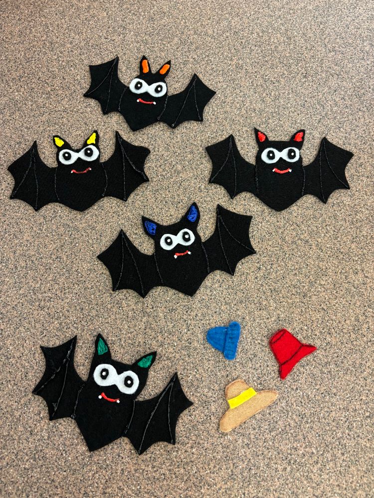 A photo of five felt bats (each with a different ear color and three tiny hats. The hats go behind the bats and the storytime kids have to guess which bat each hat is under.