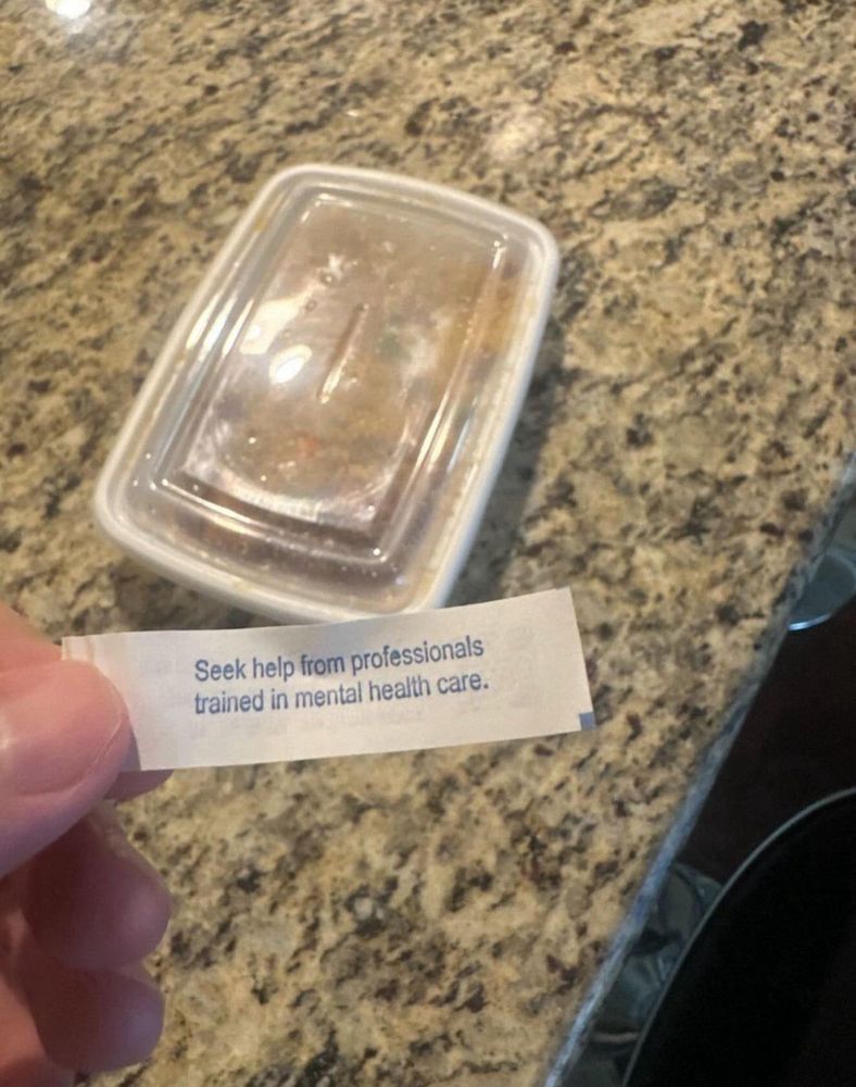 A photo of a tabletop with a plastic takeout container in the background and a hand holding a fortune in the foreground. The message on the fortune reads: Seek help from professionals trained in mental health care.