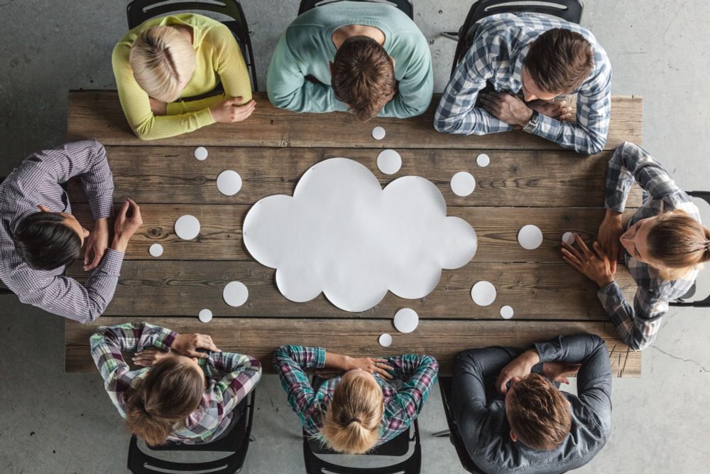 Image looking down at eight people seated around a wooden table with a blank quote bubble in the centre. 