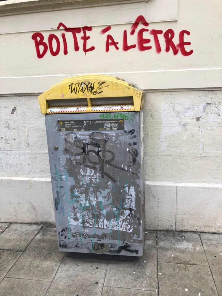 Une boîte aux lettres en ville, vieille et abîmée. Sur le mur au-dessus, quelqu’un a tagué "Boîte à l'être" (l' - e - t -r - e).