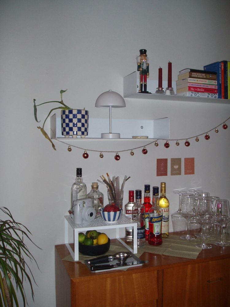 A cozy home bar setup on a mid-century wooden cabinet, decorated with a garland of small red ornaments. The shelves above hold books, candles, a nutcracker, and a checkered blue-and-white plant pot. Bottles of alcohol, glasses, and a bowl of fruit are neatly arranged below.