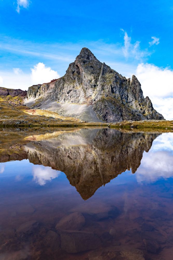 La imagen muestra una montaña rocosa de forma imponente reflejada en un lago cristalino, creando una simetría perfecta entre el paisaje y su reflejo. El cielo despejado, con nubes blancas esparcidas, resalta la claridad y viveza del entorno. Los tonos cálidos de la vegetación contrastan con las piedras grises de la montaña, enmarcando un lugar sereno y majestuoso.

