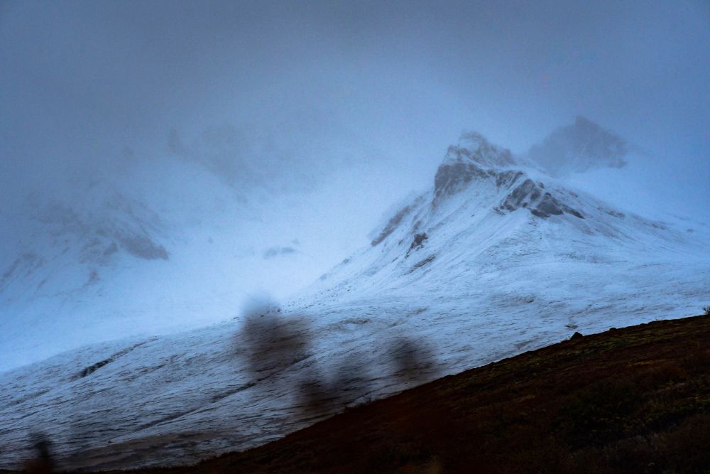 La imagen muestra un paisaje de montaña cubierto de nieve en Islandia, bajo un cielo nublado y brumoso que crea una atmósfera fría y misteriosa. Las cumbres apenas son visibles a través de la neblina, mientras que el terreno en primer plano presenta un leve contraste con tonos más oscuros, acentuando el clima inhóspito del lugar.

