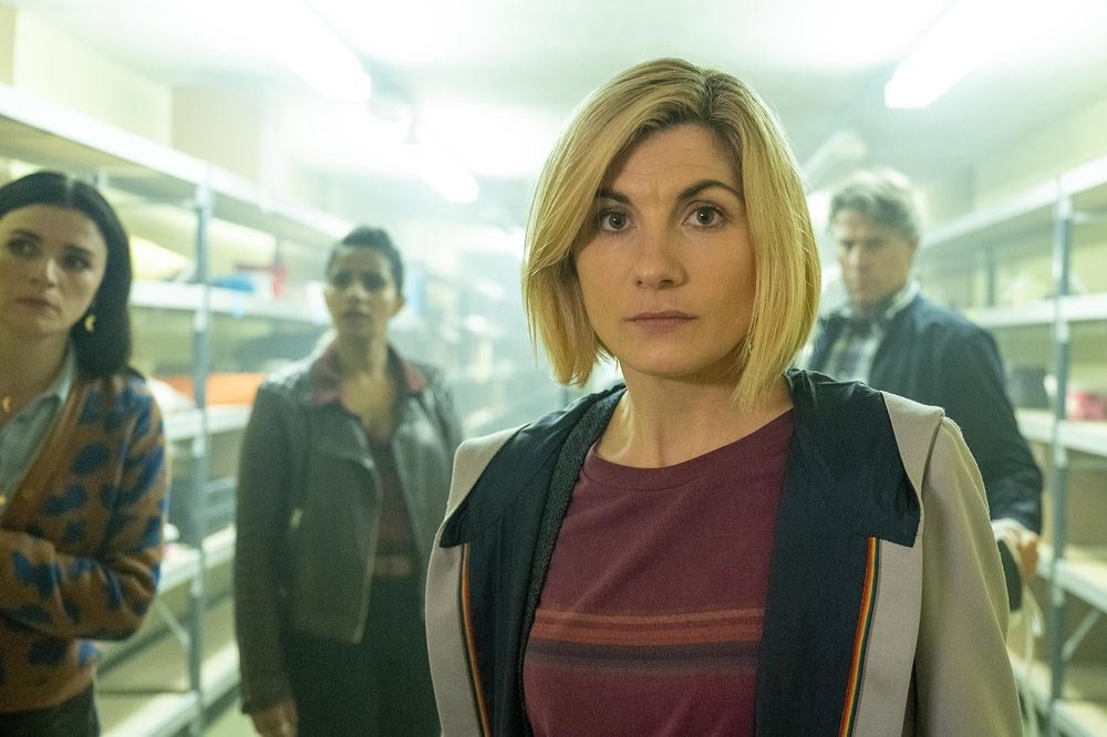 Screenshot of the Doctor Who episode Eve of the Daleks, featuring the Thirteenth Doctor at the front, and from left to right Sarah, Yaz and Dan, all standing in a room full of shelves with diverse objects.