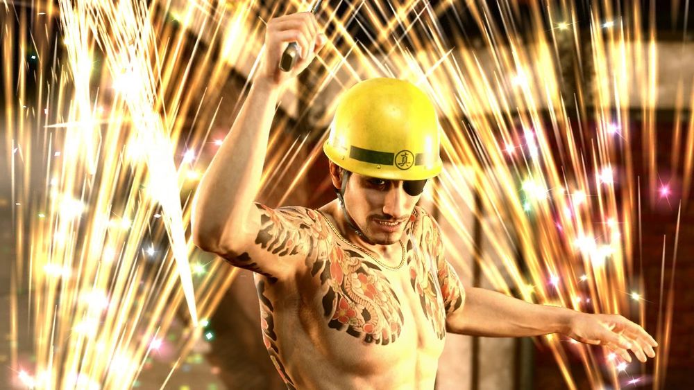 Captura de Yakuza Kiwami 2 donde aparece Goro Majima sin camisa, con todos sus tatuajes al aire, con un casco de obra amarillo y fuegos artificiales detrás. Aparece con el brazo derecho en un ángulo de 90 grados sujetando su cuchillo, y en posición de pelea. 
